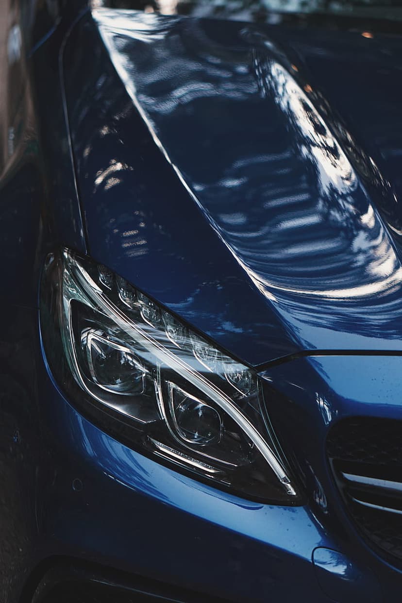 Dark blue car headlight showing detailed lens housing and glossy reflections of trees on the vehicle hood.