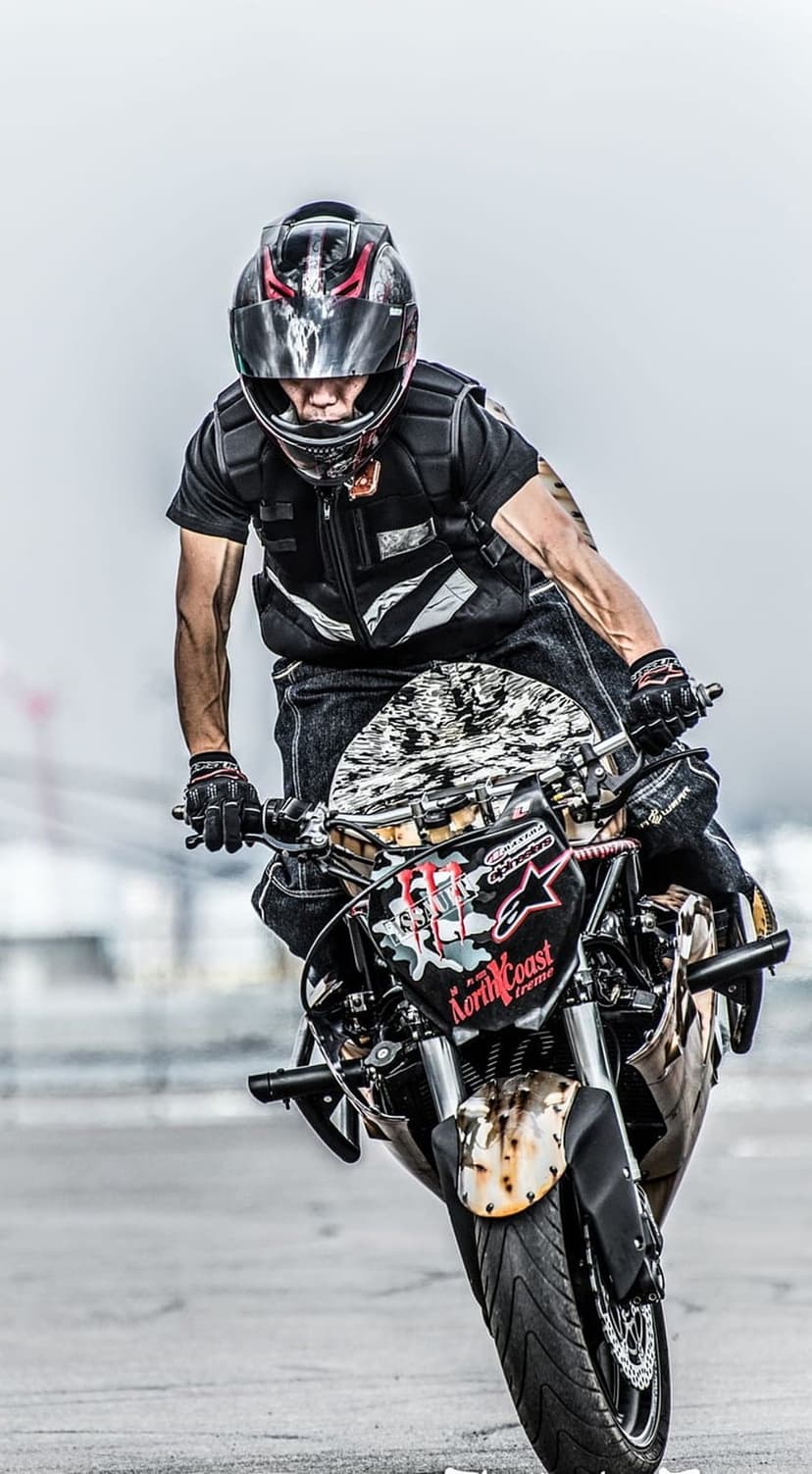 Motorcycle stunt rider wearing a black helmet and protective gear performs a low-angled wheelie on an urban street.