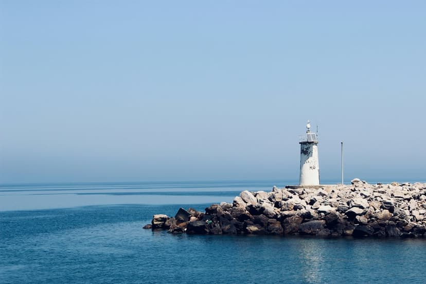 White lighthouse on a rocky jetty beside a calm blue sea under a cloudless sky during a bright day.