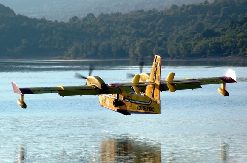 Yellow Canadair CL-415 firefighting seaplane flying low over a forest lake against green hills and blue sky.