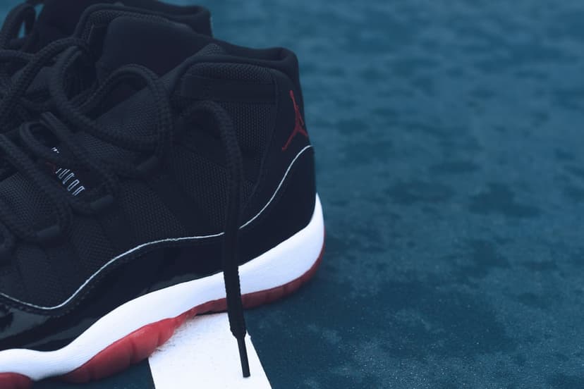 Jordan 11 Bred sneakers with black patent leather and red translucent soles rest against a blue backdrop.