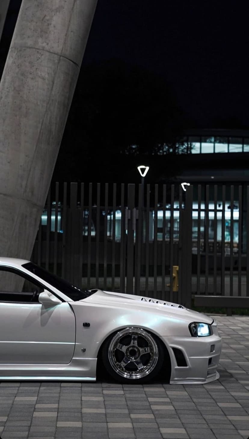White modified sports car with chrome rims parked beside a concrete pillar under urban city lights at night.