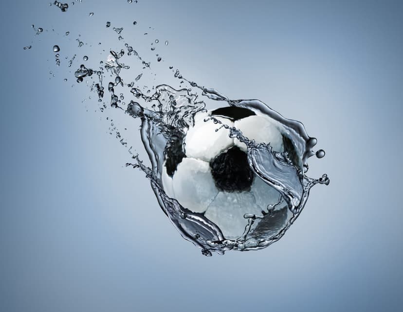 Soccer ball bursting through a splash of clear water droplets against a soft blue gradient background.