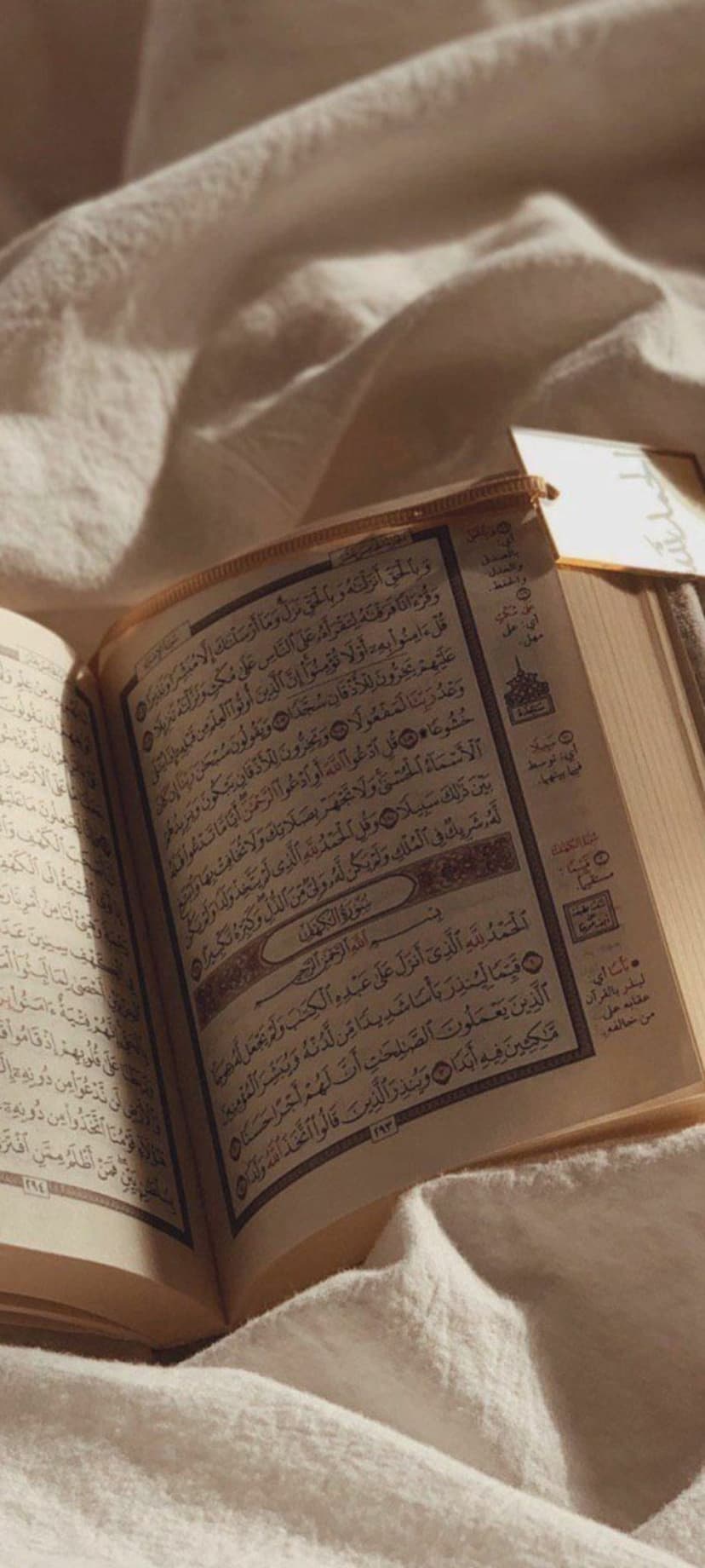 Open Quran with Arabic script rests on folded cream cloth under warm golden sunlight streaks in a vertical frame.