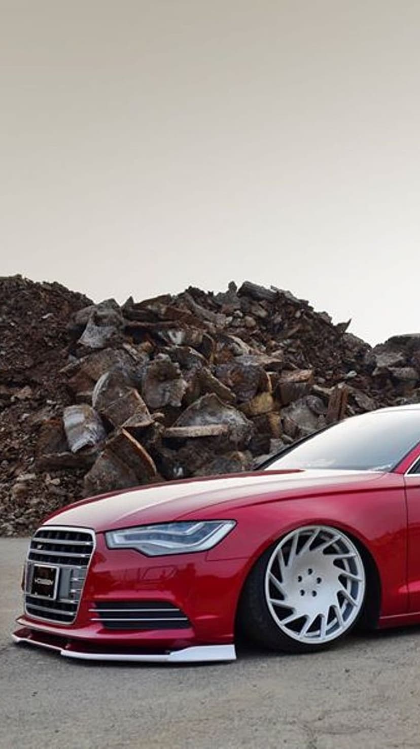 Red Audi A6 sedan with slammed suspension and white multi-spoke wheels parked on a bed of grey crushed stones.