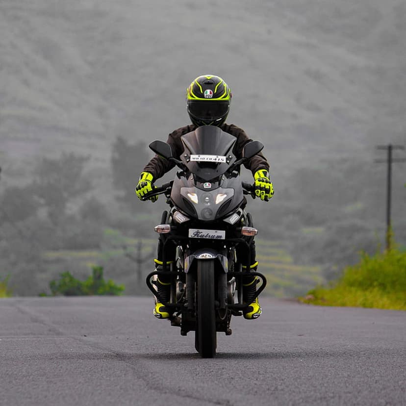 Motorcyclist in yellow gloves rides a black bike on a grey asphalt road through dense morning fog and hills.