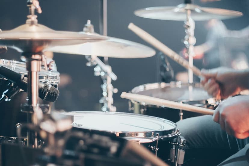 Drummer's hands holding wooden sticks strike a metallic cymbal and snare drum under bright stage lights.