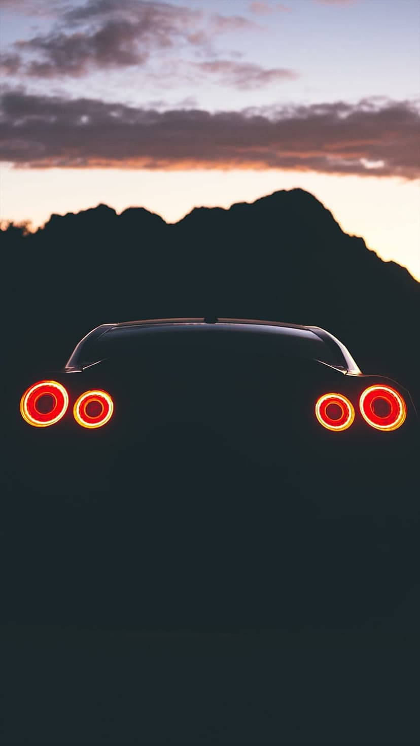 Red circular taillights on a dark car face silhouetted mountain peaks under a colorful twilight sky.