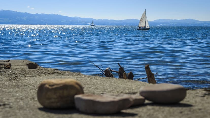 Sailboat on a sparkling sapphire lake with distant mountain peaks, driftwood, and rocks along the shore.