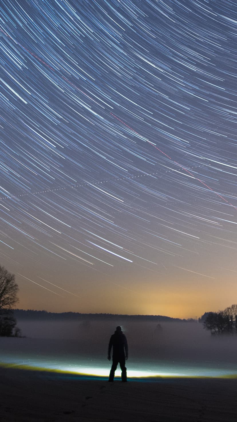 Solitary person gazes at circular star trails arching over a glowing horizon in a deep blue nocturnal landscape.