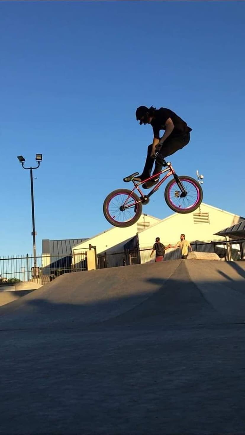 BMX rider in silhouette jumping a red and blue bike over a skatepark against a bright, clear blue sky.