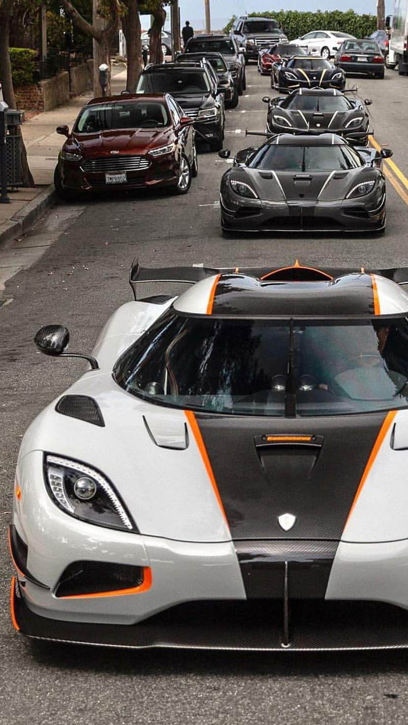 Koenigsegg hypercars with orange details parked in a row on a sun-drenched street under green leafy trees.