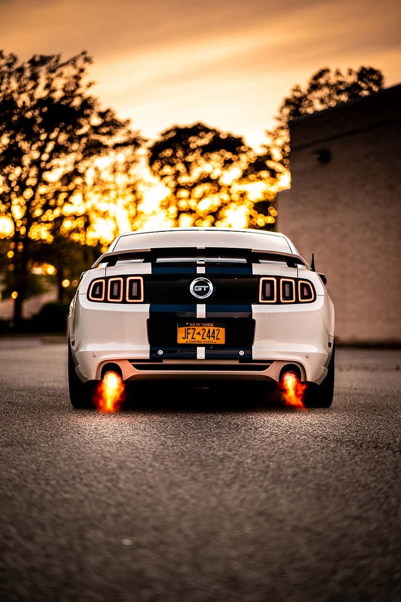 White Ford Mustang GT with blue stripes emits orange exhaust flames on asphalt under a warm sunset sky.