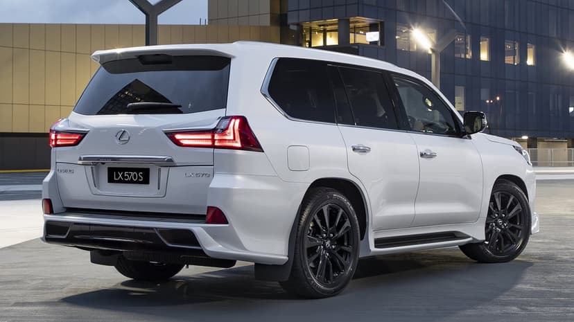 White Lexus LX 570 S SUV with glowing red taillights and black wheels parked in front of a modern glass building.