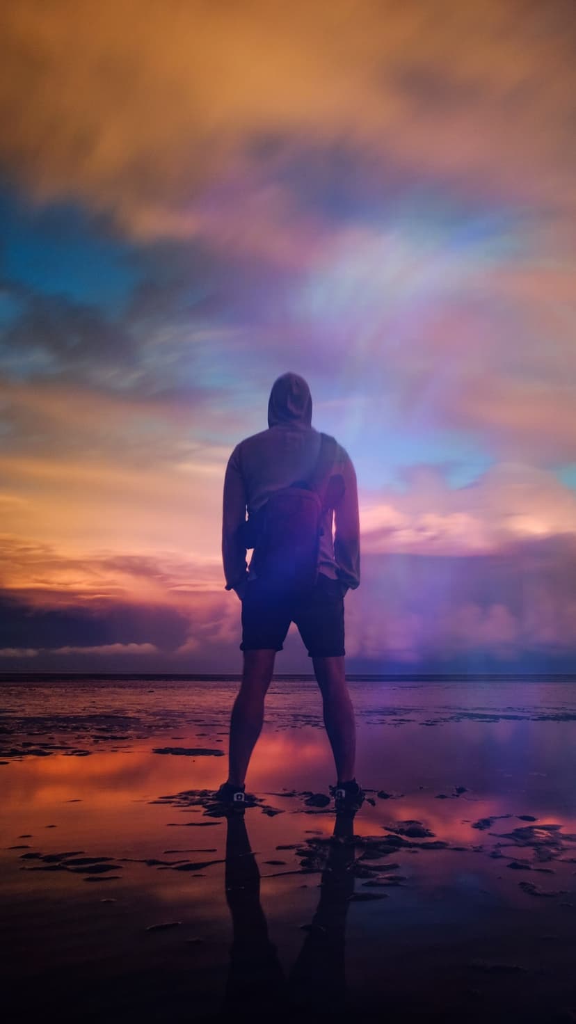 Person standing on reflective wet ground gazing at a vibrant orange and pink sunset sky during twilight hours.