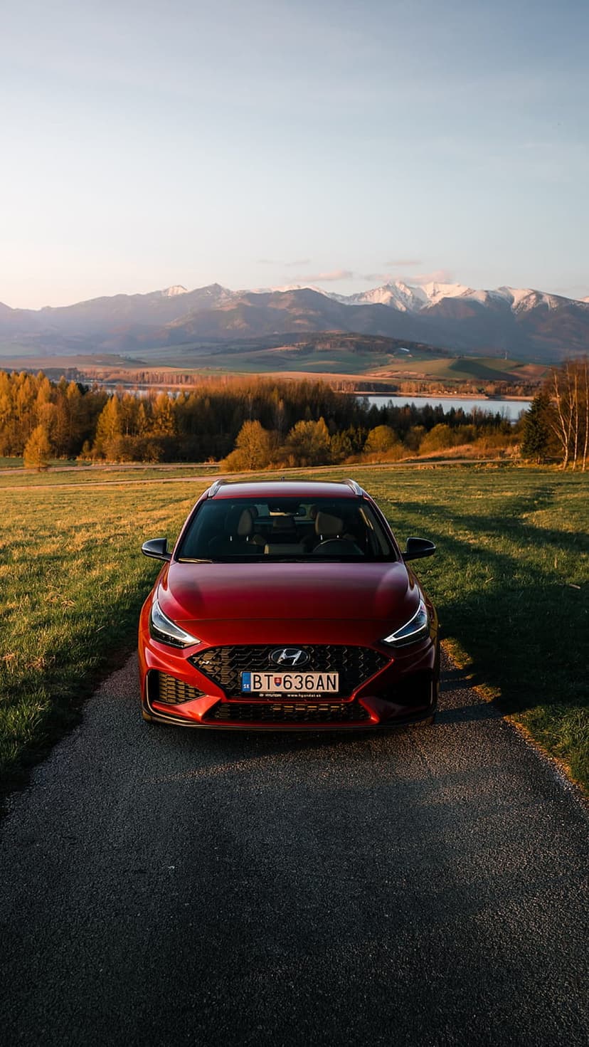 Red Hyundai hatchback parked on an asphalt mountain road with snowy peaks and autumn trees in the background.