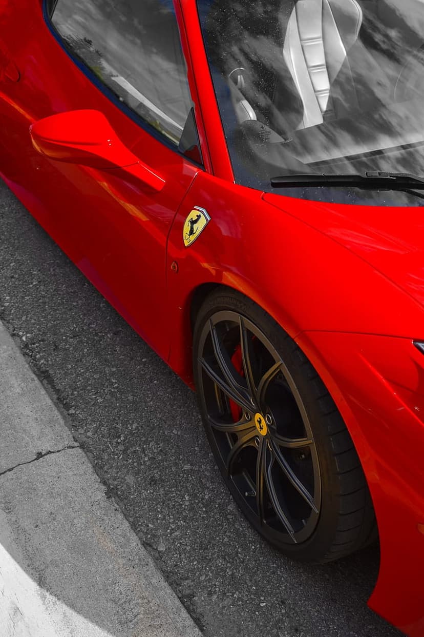 Ferrari prancing horse emblem on a glossy red car door above a black multi-spoke wheel and dark gray asphalt.