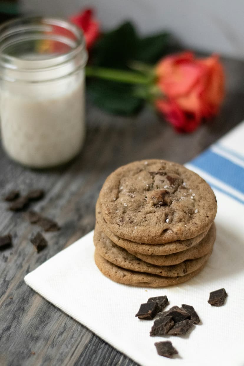Stacked chocolate chip cookies with sea salt grains beside red roses and a glass of milk on a white napkin.