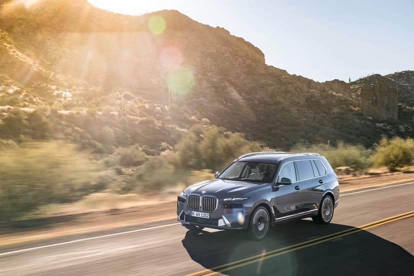 Dark gray BMW SUV driving on a desert highway during golden hour with mountains and sun flare in the background.