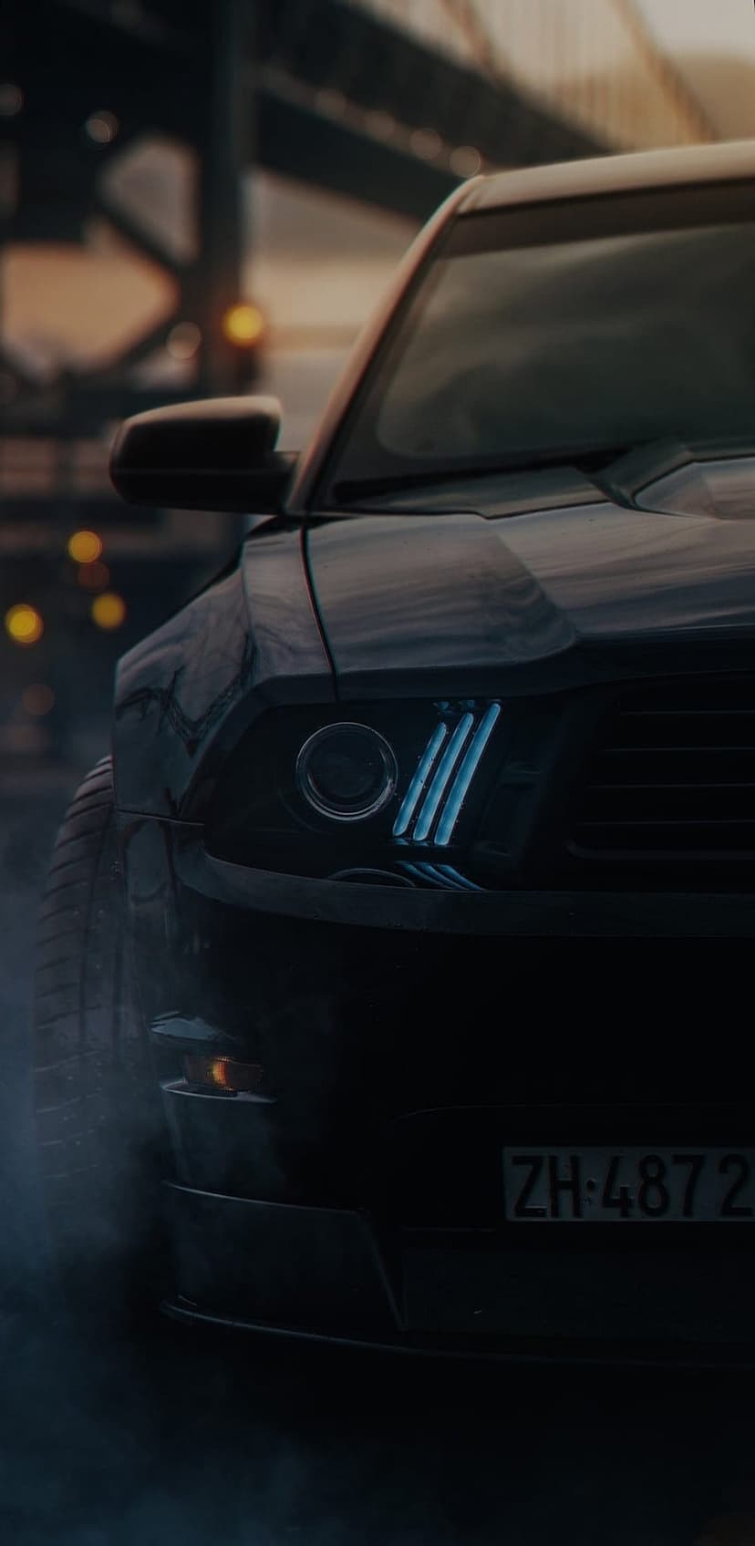 Ford Mustang with blue LED headlights parked in smoke with a Swiss license plate under city night lights.