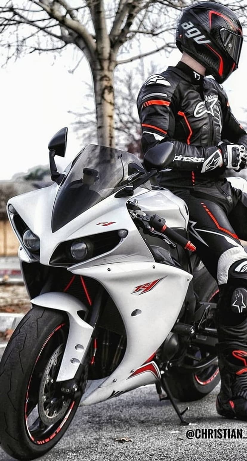 Motorcyclist in red and black racing gear sits on a white Yamaha R1 sportbike on asphalt near a bare tree.