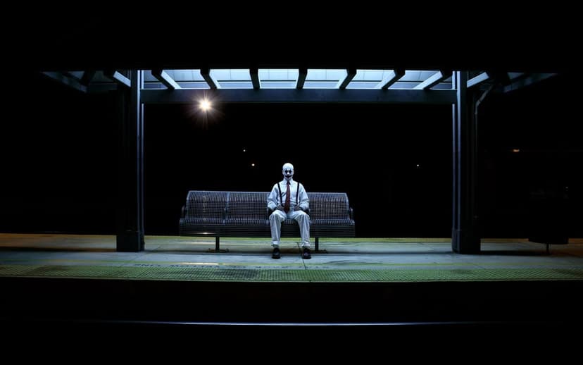 Man in clown makeup wearing a suit sits on a bench under glowing bus stop lights against a black night sky.