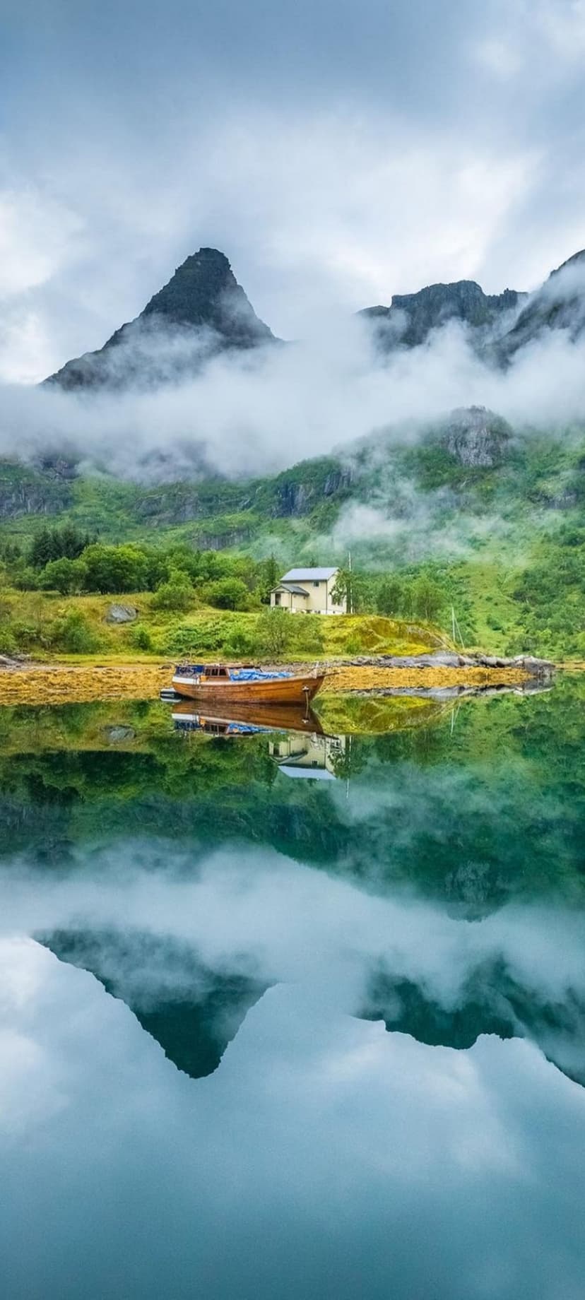Jagged mountains shrouded in mist reflect in a green fjord next to a wooden boat and a small house on the shore.