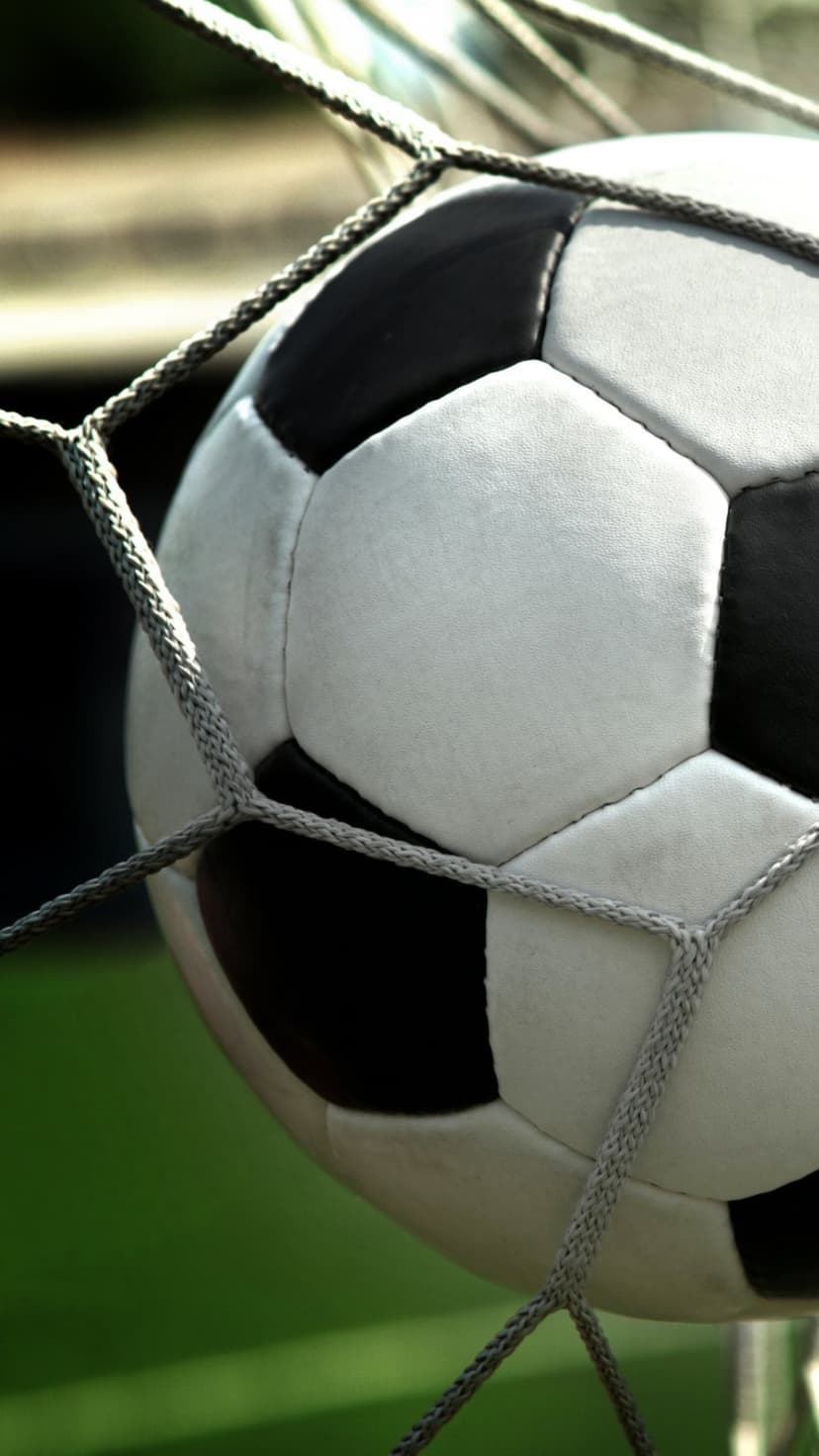 Soccer ball bulging into white goal netting with a blurred green grass stadium background in a close-up shot.