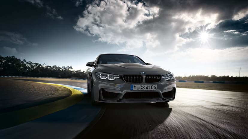 BMW M3 CS driving on a race track curve with motion blur, blue and yellow curbing, and a dramatic cloudy sunburst sky.