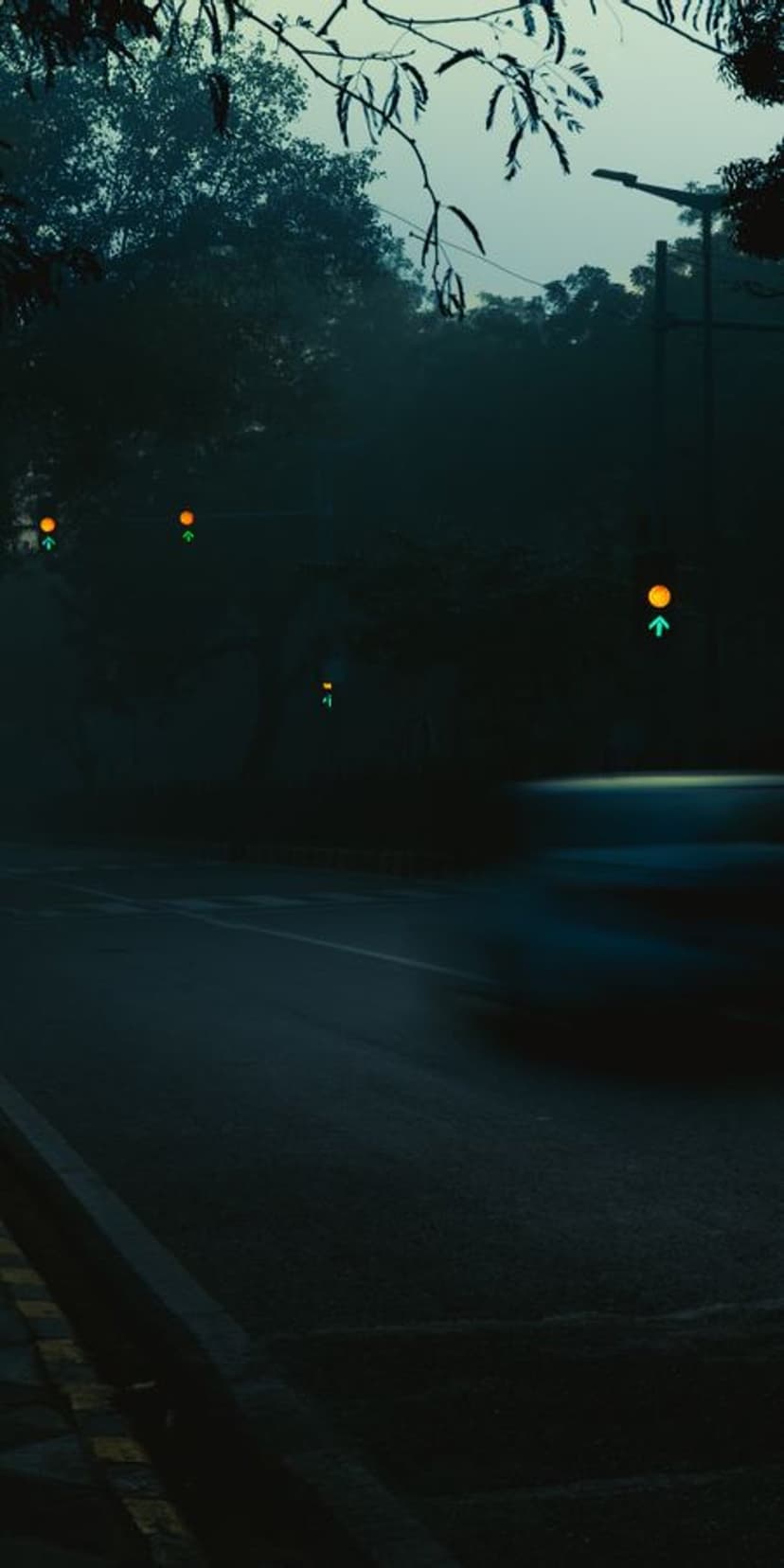 Blurry car moving along a dark foggy street at night with glowing orange streetlights and green traffic signals.