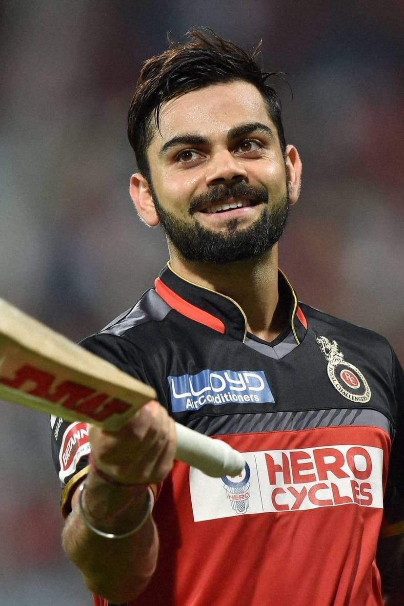 Virat Kohli smiles while holding a cricket bat, wearing a red and black Royal Challengers Bangalore team jersey.