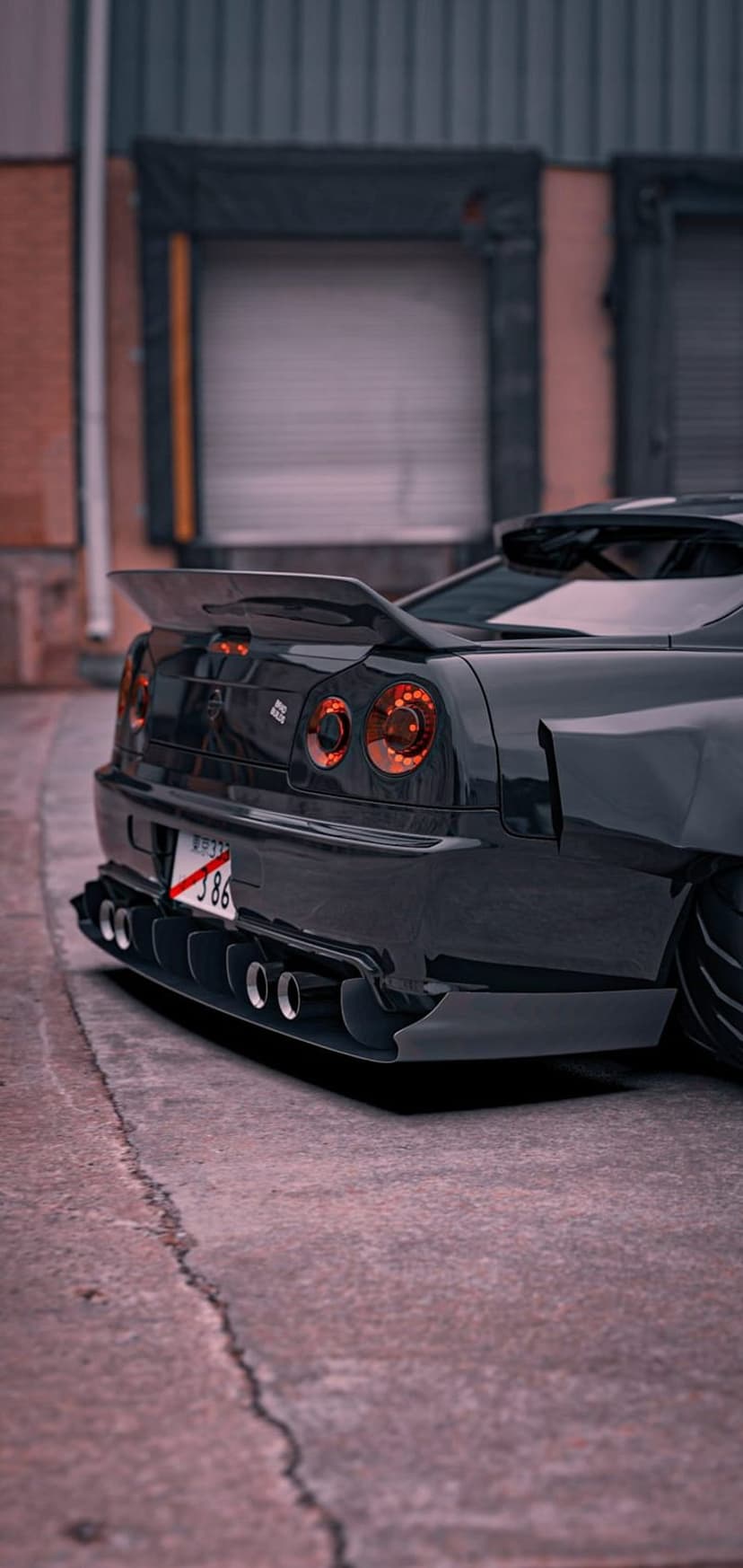 Black Nissan Skyline R34 rear view featuring a large wing and quad exhausts on cracked industrial asphalt.