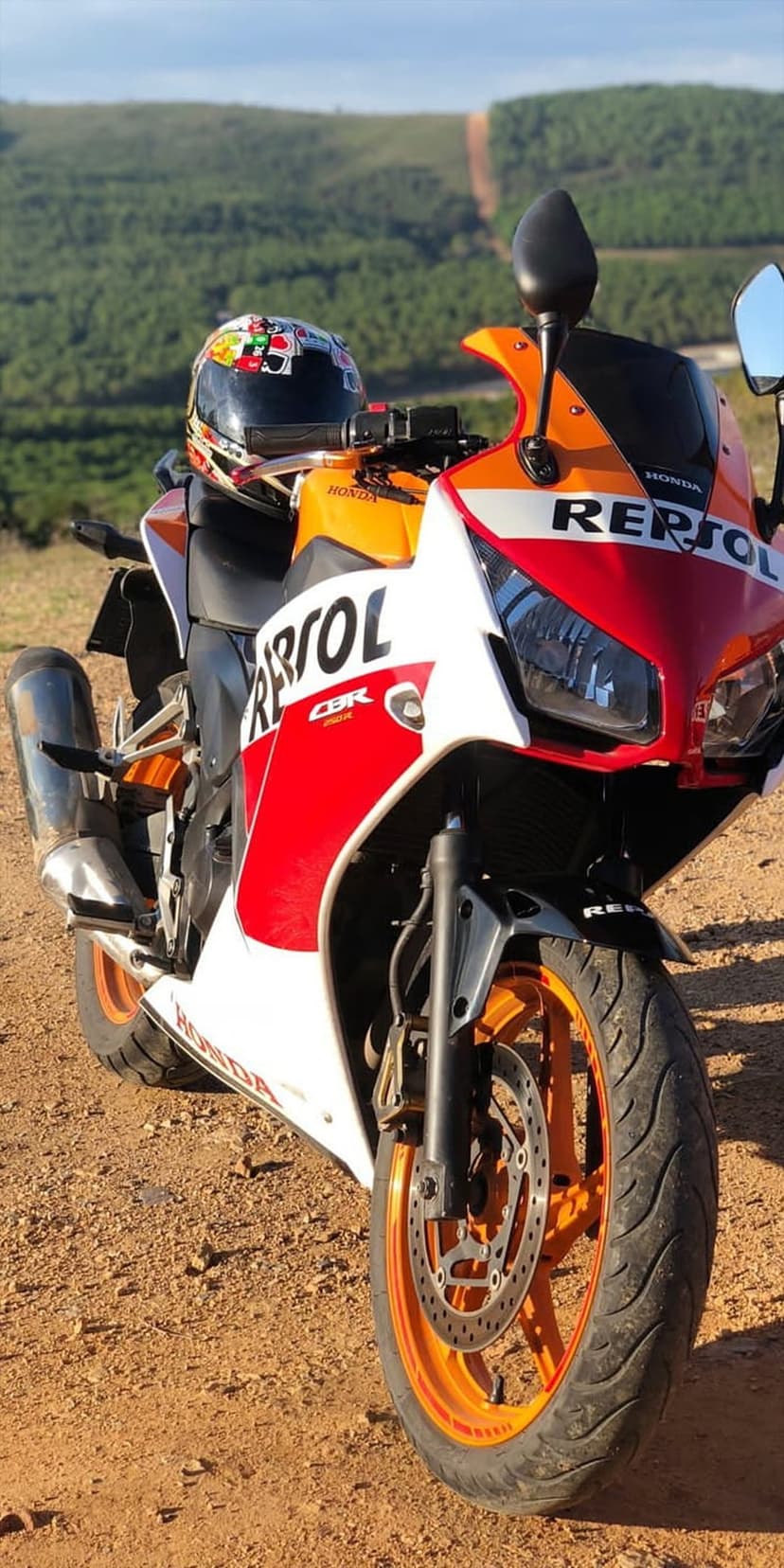 Repsol Honda CBR motorcycle with a helmet on the seat parked on a grassy hillside overlooking distant green mountains.