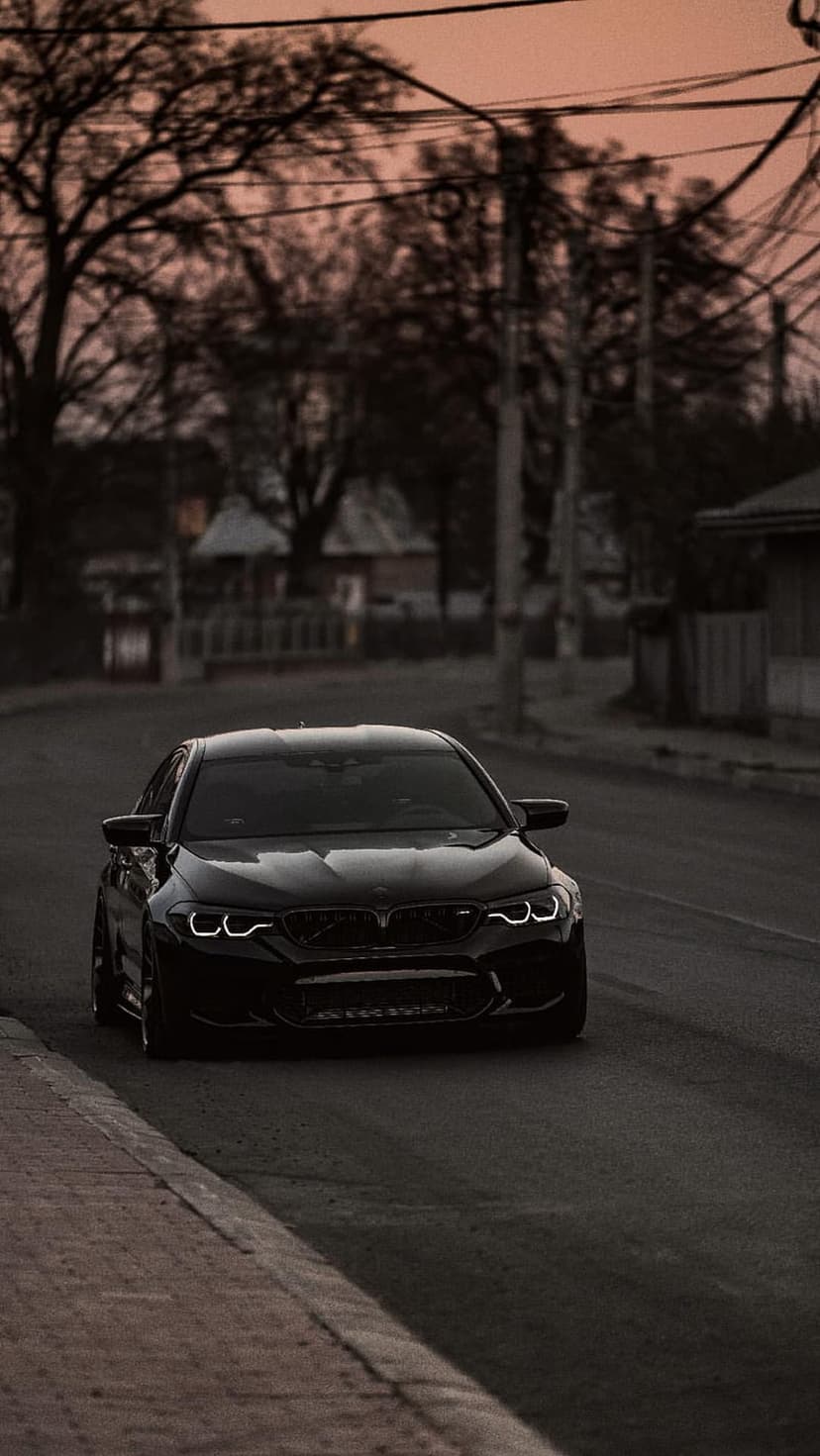 Black BMW M5 sedan with glowing headlights on a residential street under a hazy orange sunset sky with bare trees.