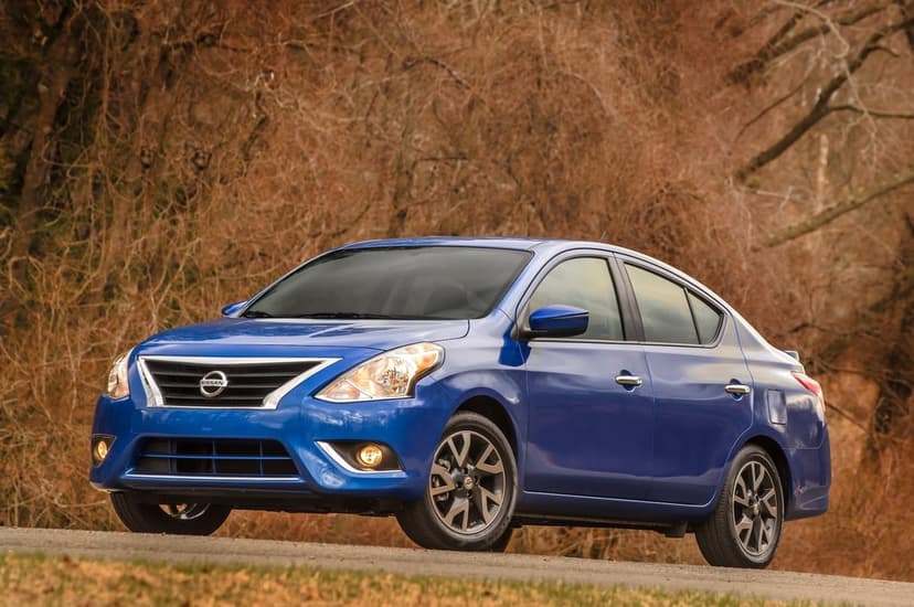 Blue Nissan Versa sedan parked on a road with a background of blurred brown autumn trees in daylight.