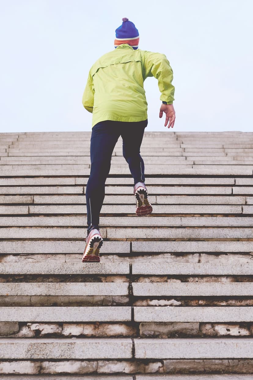Runner in a lime green jacket and black leggings climbs outdoor concrete stairs against a bright overcast sky.