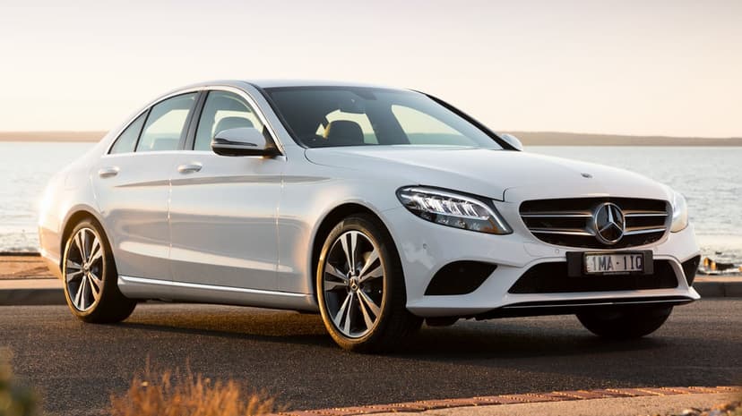 White Mercedes-Benz sedan parked on a coastal road at sunset with calm water in the background.