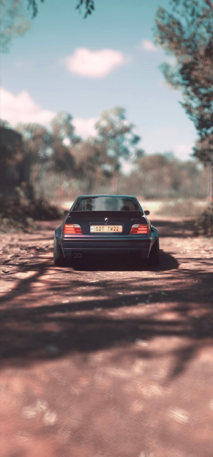 BMW E36 M3 with a widebody kit drives on a dusty dirt road through a sunny forest under a clear blue sky.
