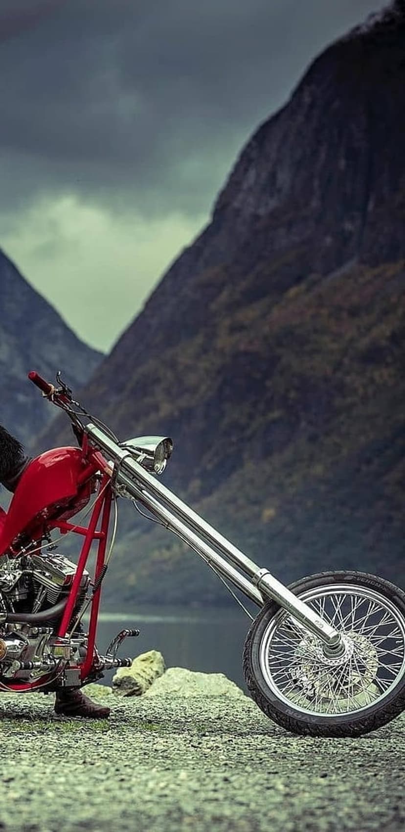 Red custom chopper motorcycle parked on a rocky beach with towering mountains and a cloudy sky in the background.