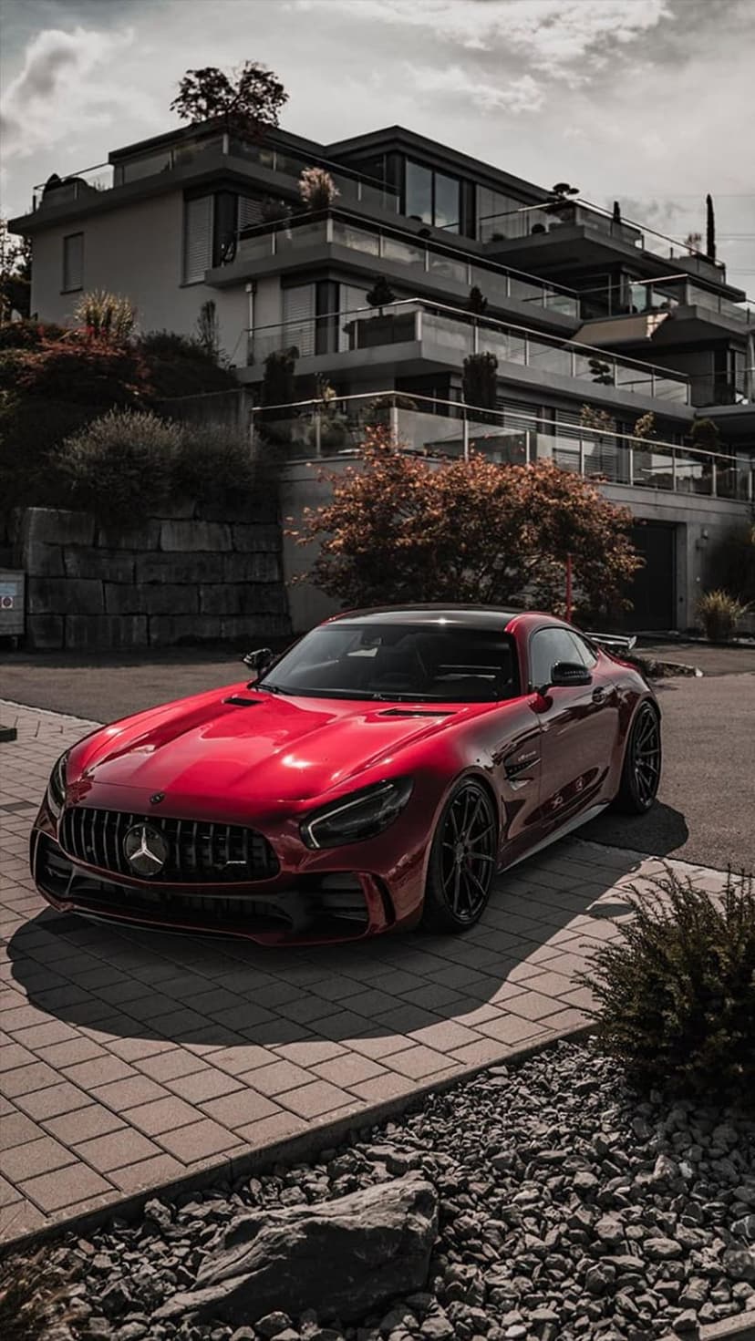 Red Mercedes-AMG GT parked on a stone driveway in front of a modern glass villa with lush green landscaping.
