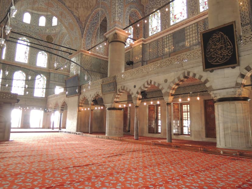 Ornate red carpets lead to massive columns and blue tiled arches under stained glass in the Blue Mosque.