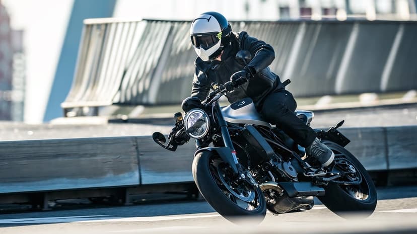 Motorcyclist in a leather jacket leans a silver Husqvarna Vitpilen 701 into a sharp turn on an urban bridge.