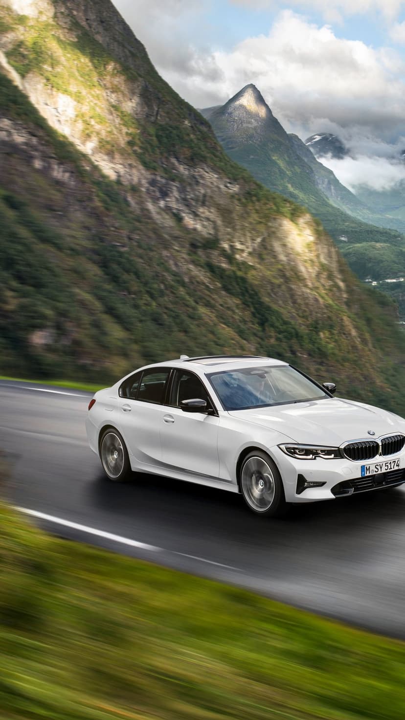 White luxury sedan driving on a wet mountain road with steep green cliffs and clouds in the background.