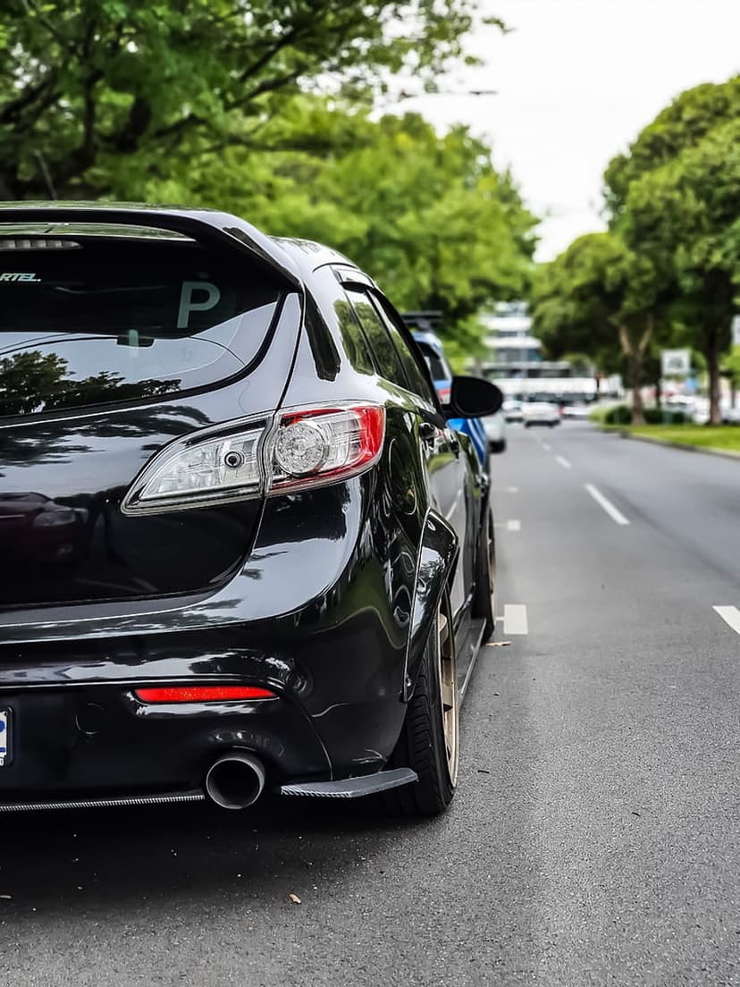 Black Mazda 3 hatchback featuring widebody fenders and custom wheels parked on a city street with green leafy trees.