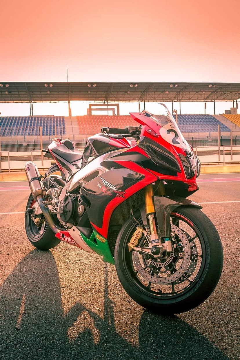 Red and black sportbike parked on a tarmac race track during a golden sunset with stadium seating behind.