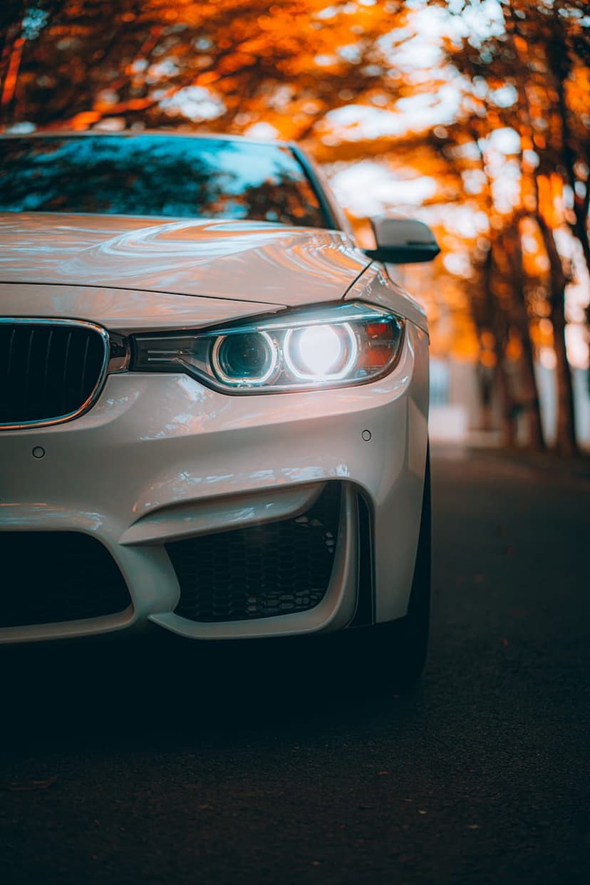 White BMW M Sport car with glowing halo headlights on an asphalt road lined with orange autumn forest trees.