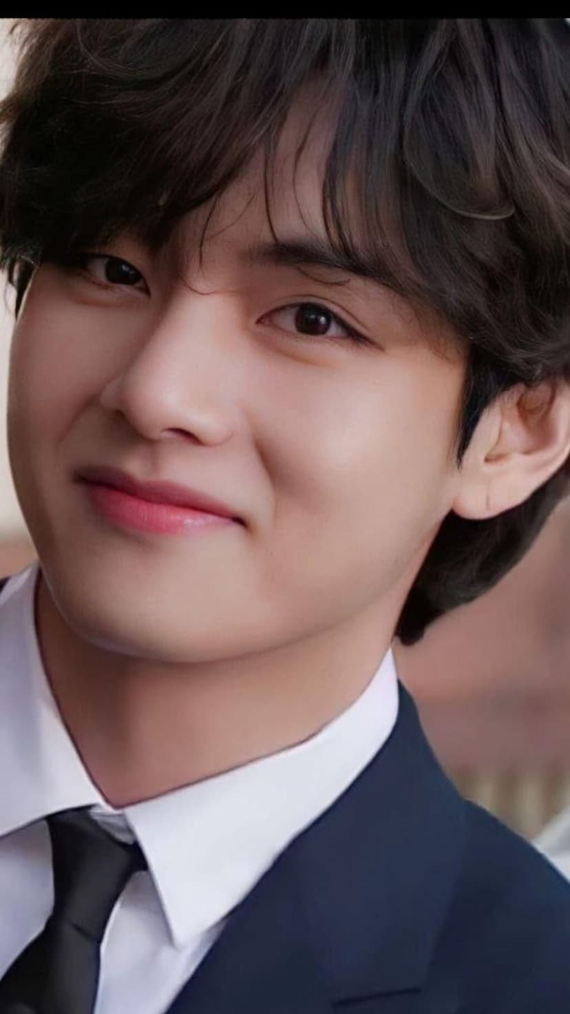 Kim Taehyung smiles gently while wearing a black suit and tie, featuring soft wavy hair in a close-up portrait.