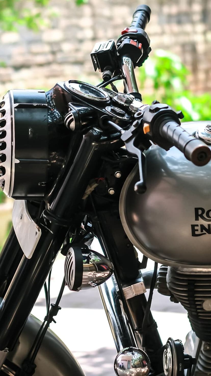 Royal Enfield motorcycle front featuring a grilled headlight, silver fuel tank, and blurred garden background.