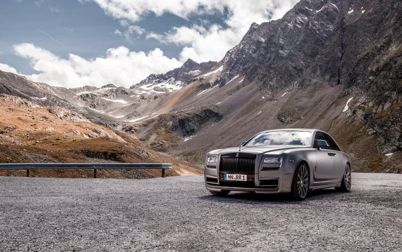 Matte grey Rolls-Royce parked on a gravel mountain road with rocky peaks and a cloudy blue sky behind it.