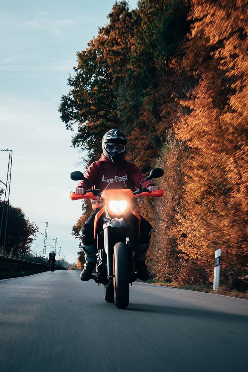Motorcyclist on an orange bike rides through orange autumn trees with a bright headlight shining on the asphalt.